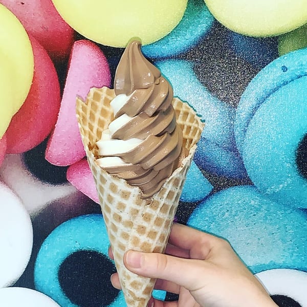 Hand holding a waffle cone with swirled chocolate and vanilla soft-serve ice cream against a colorful background.