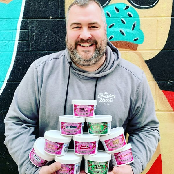 Smiling man in a grey hoodie holding a stack of various Chocolate Moose ice cream pints.